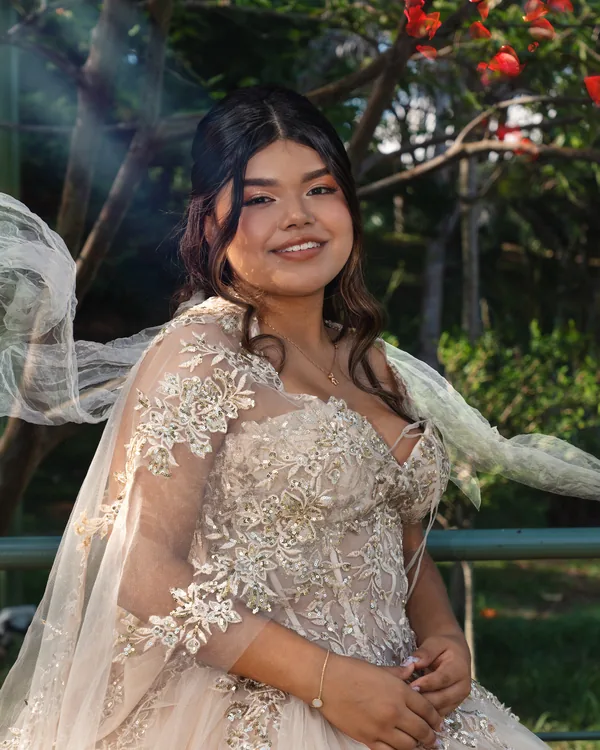 Quinceañera sonriente con vestido champaña bordado en dorado y el velo al viento entre árboles