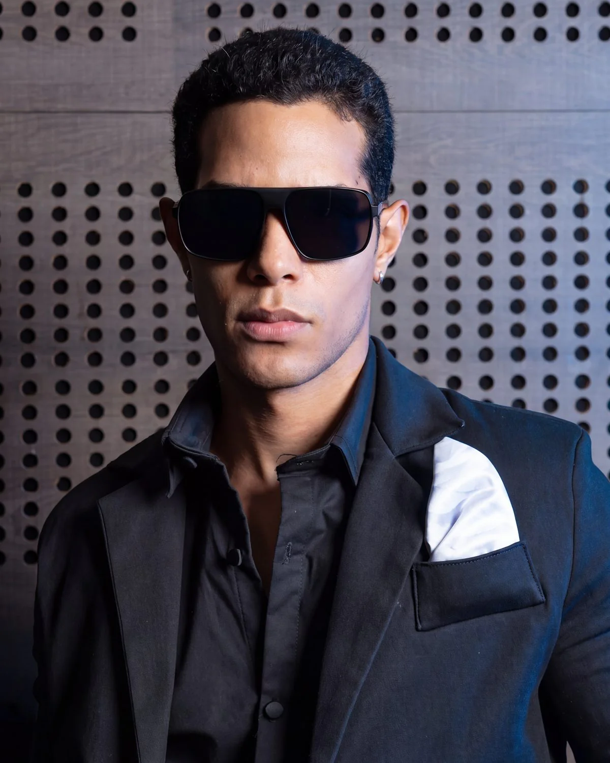Modelo con gafas de sol y traje negro frente a un panel acústico perforado