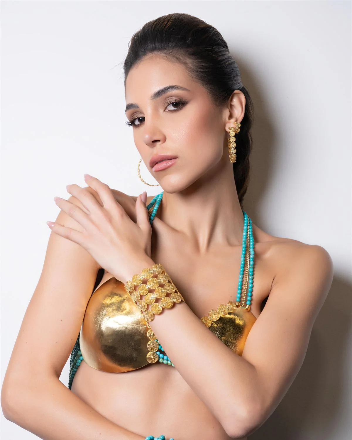 Modelo con pectoral de latón dorado, brazalete de filigrana y collar de turquesas sobre fondo blanco de estudio